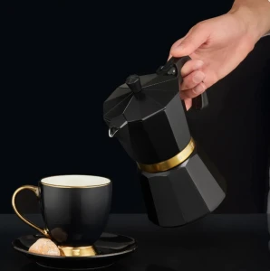 Czarna kawiarka ciśnieniowa ze złotym detalem trzymana w dłoni nad elegancką czarno-złotą filiżanką do espresso.