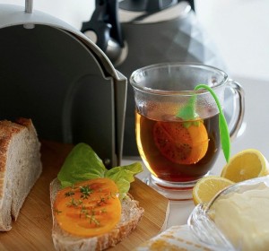 Szklany kubek napełniony herbatą z zaparzaczem w kształcie cytryny. Obok znajduje się kanapka z sałatą i pomidorem. W pobliżu otwarty szary chlebak a za nim szary czajnik.