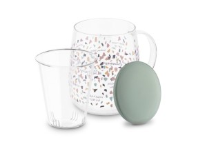 Szklany kubek z kolorową grafiką o który opiera się ceramiczna zielona pokrywka. Przed kubkiem wyciągnięty szklany zaparzacz.