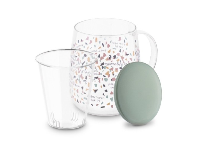 Szklany kubek z kolorową grafiką o który opiera się ceramiczna zielona pokrywka. Przed kubkiem wyciągnięty szklany zaparzacz.